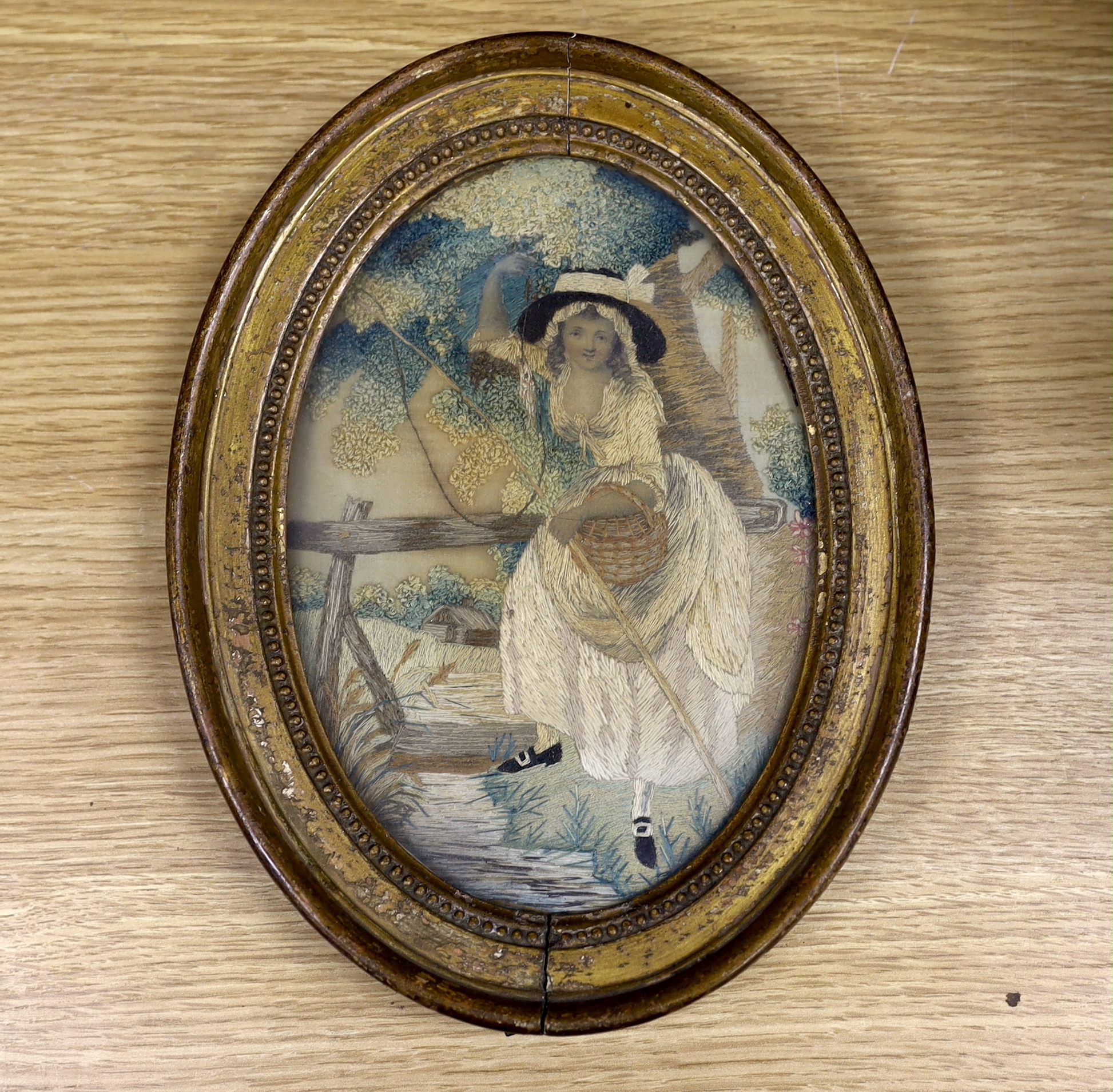 An early George IV oval silk work needlework pastoral embroidery, of a young girl with a fishing rod and basket under a tree by a bridge and stream, worked in fine wools and silks in stem stitch and a large knotting stit
